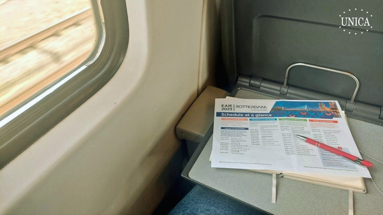 Paper, notebook and pen displayed on a table on a train seat. Snippet of the train's window on the left side
